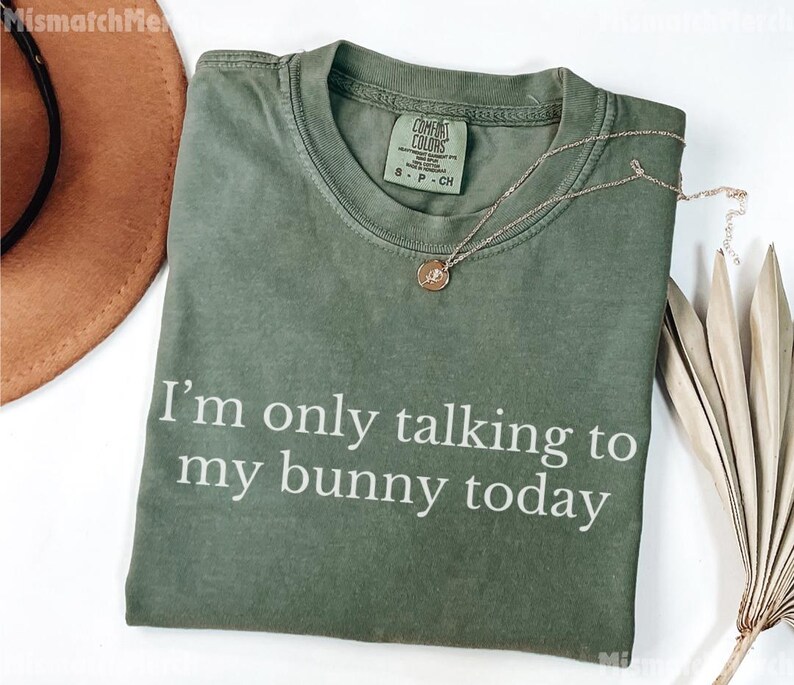 May include: Olive green t-shirt with the text "I'm only talking to my bunny today" in white. A gold necklace with a pendant and a brown hat are also visible. The shirt is a Comfort Colors brand.