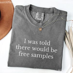 May include: A gray Comfort Colors t-shirt with the text "I was told there would be free samples" in white. A gold necklace with a pendant is on the shirt. A brown hat and dried plant are in the background.
