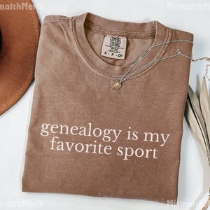 May include: A brown t-shirt with the text "genealogy is my favorite sport" printed in white. The shirt is folded and laid flat on a surface.