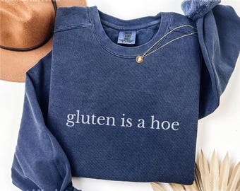 May include: A navy blue crew neck sweatshirt with the phrase "gluten is a hoe" in white lettering. A gold necklace with a small pendant is draped on the sweatshirt. A brown hat is visible in the background.