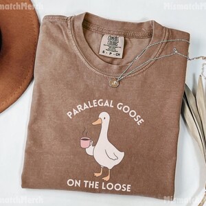 May include: A brown Comfort Colors t-shirt featuring the text "PARALEGAL GOOSE ON THE LOOSE" and a cartoon goose with a coffee cup. A silver necklace with a pendant is draped across the shirt.