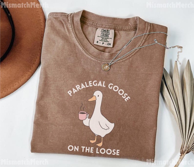 May include: A brown Comfort Colors t-shirt featuring the text "PARALEGAL GOOSE ON THE LOOSE" and a cartoon goose with a coffee cup. A silver necklace with a pendant is draped across the shirt.