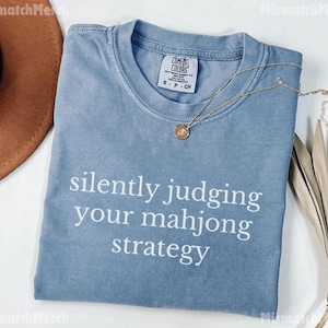 May include: A light blue t-shirt with the text "silently judging your mahjong strategy" in white. The shirt has a crew neck and short sleeves. A gold necklace with a pendant is draped across the shirt.