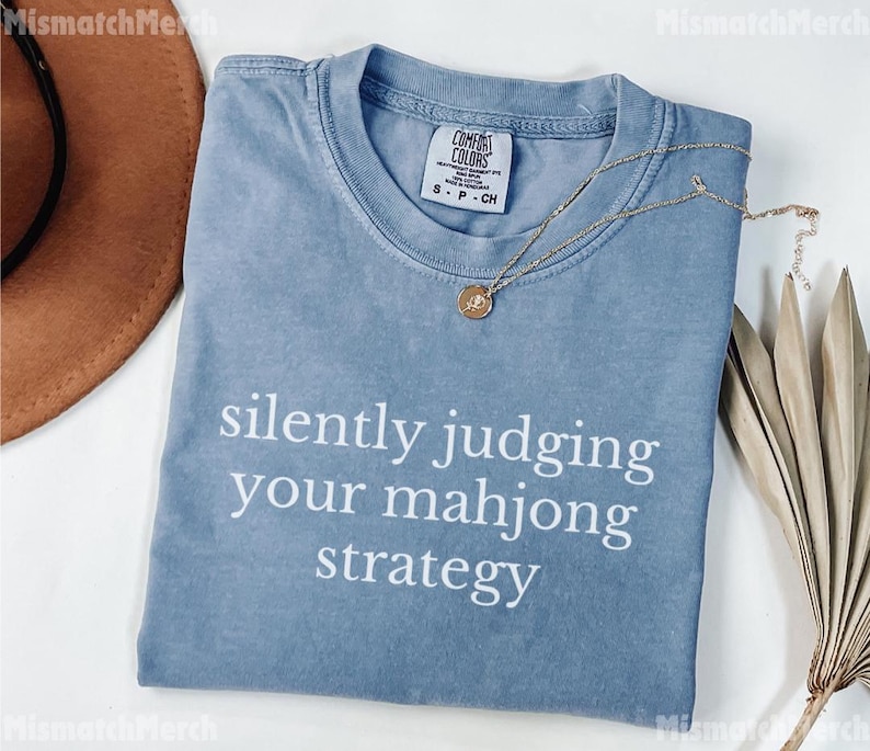 May include: A light blue t-shirt with the text "silently judging your mahjong strategy" in white. The shirt has a crew neck and short sleeves. A gold necklace with a pendant is draped across the shirt.