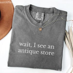 May include: A gray t-shirt with the text "wait, I see an antique store" in white. The shirt has a crew neck and a small tag near the collar. A silver necklace with a pendant is draped across the shirt.