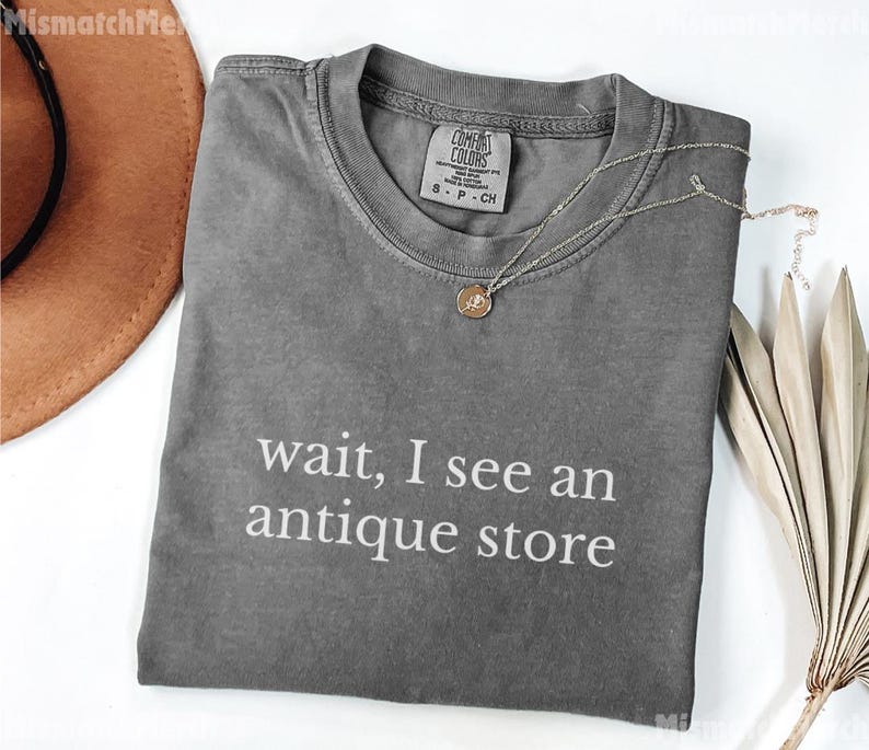 May include: A gray t-shirt with the text "wait, I see an antique store" in white. The shirt has a crew neck and a small tag near the collar. A silver necklace with a pendant is draped across the shirt.