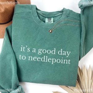 May include: A teal green sweatshirt with the phrase "it's a good day to needlepoint" in white lettering. A gold necklace with a small pendant is draped on the front. A brown hat is visible in the upper left corner.