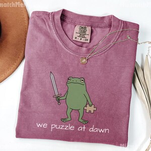 May include: A mauve crewneck t-shirt featuring a green frog illustration holding a silver sword and a tan puzzle piece. The text below the frog reads, we puzzle at dawn. The shirt has a Comfort Colors brand label on the inside collar area.