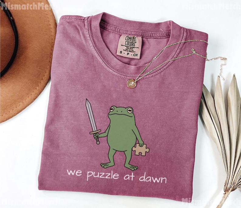 May include: A mauve crewneck t-shirt featuring a green frog illustration holding a silver sword and a tan puzzle piece. The text below the frog reads, we puzzle at dawn. The shirt has a Comfort Colors brand label on the inside collar area.