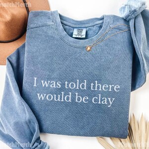May include: A blue crew neck sweatshirt with the text "I was told there would be clay" in white. A gold necklace with a pendant is draped across the front. A brown hat is visible in the upper left corner.