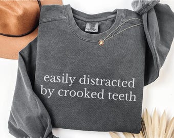 Może przedstawiać: Ciemnoszara bluza z długim rękawem z białym napisem "easily distracted by crooked teeth". Złoty naszyjnik z wisiorkiem jest zarzucony na koszulę. Brązowy kapelusz jest widoczny w lewym górnym rogu.