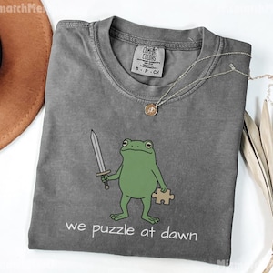 May include: A charcoal gray short-sleeve t-shirt featuring a graphic of a green frog holding a silver sword and a tan puzzle piece. Below the frog, the text reads "we puzzle at dawn" in white. The shirt has a crew neckline and a Comfort Colors label.