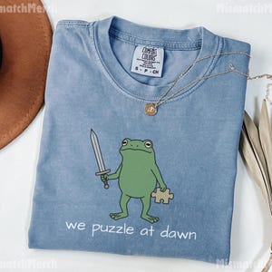 May include: A blue short-sleeved t-shirt featuring a green frog illustration holding a silver sword and a tan puzzle piece. The text below the frog reads "we puzzle at dawn". The shirt has a Comfort Colors brand label on the inside neck area.