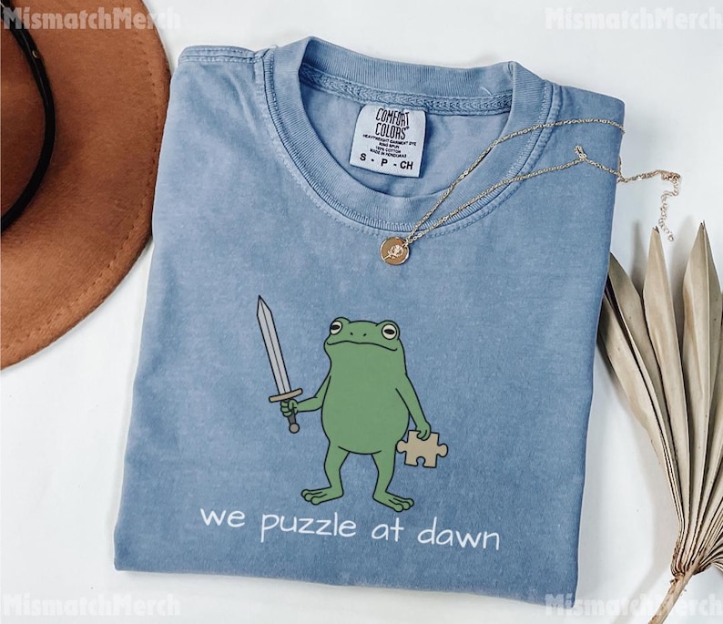 May include: A blue short-sleeved t-shirt featuring a green frog illustration holding a silver sword and a tan puzzle piece. The text below the frog reads "we puzzle at dawn". The shirt has a Comfort Colors brand label on the inside neck area.