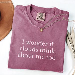 May include: A dusty rose t-shirt with the text "I wonder if clouds think about me too" in white. The shirt has a crew neck and a small tag. A gold necklace with a pendant is draped across the shirt.