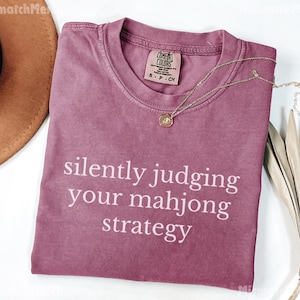 May include: A dusty rose Comfort Colors t-shirt with the phrase "silently judging your mahjong strategy" in white. A gold necklace with a pendant rests on the shirt. A brown hat and dried plant are in the background.