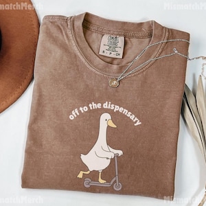 May include: Brown t-shirt with the phrase "off to the dispensary" above a cartoon duck riding a scooter. The shirt has a crew neck and short sleeves. A silver necklace with a pendant is on the shirt.