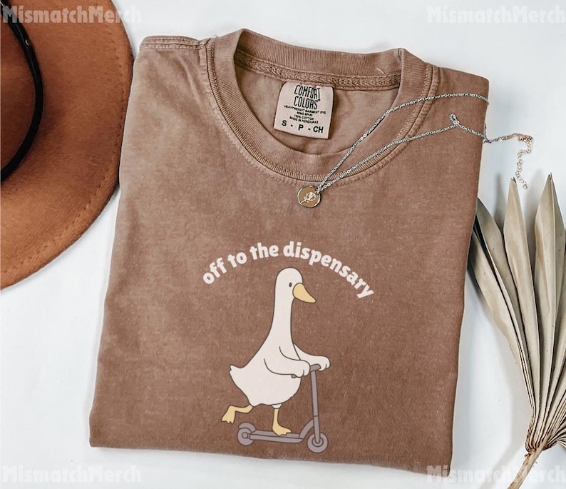 May include: Brown t-shirt with the phrase "off to the dispensary" above a cartoon duck riding a scooter. The shirt has a crew neck and short sleeves. A silver necklace with a pendant is on the shirt.
