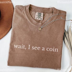May include: A brown t-shirt with the phrase "wait, I see a coin" printed in white. A silver necklace with a coin pendant rests on the shirt. The shirt has a Comfort Colors tag.