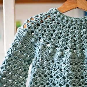Puede incluir: Un vestido de beb&eacute; de ganchillo azul claro con un delicado patr&oacute;n de calado y peque&ntilde;as bolitas de ganchillo. El vestido tiene un escote redondo y mangas largas.