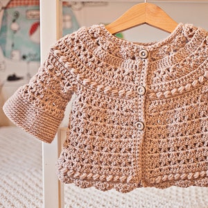 May include: A beige crocheted cardigan with a button fastening and bell sleeves.