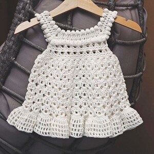 May include: A white crocheted baby dress with a scalloped hem and a delicate pattern. The dress is hanging on a wooden hanger.