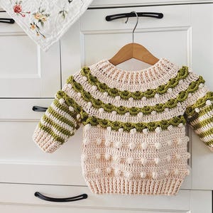 May include: A handmade, light pink and green crochet sweater for a baby. The sweater features a round neckline, long sleeves, and a textured design with bobbles and striped details. The sweater is displayed on a wooden hanger.