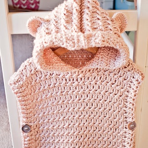 May include: A pink crocheted hooded vest with bear ears and buttons. The vest is made with a textured stitch pattern.
