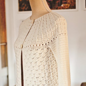 May include: A white crocheted cardigan with a button fastening. The cardigan features a delicate openwork pattern and a slightly flared shape.