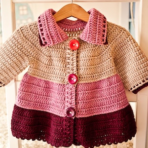 Puede incluir: Un cárdigan de bebé tejido a crochet con un esquema de color rosa, beige y burdeos. El cárdigan tiene un cuello y botones en la parte delantera.