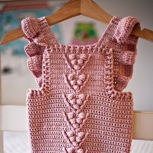 May include: A pink crocheted baby onesie with a ruffled neckline and a decorative stitch pattern. The onesie has a button closure at the bottom.