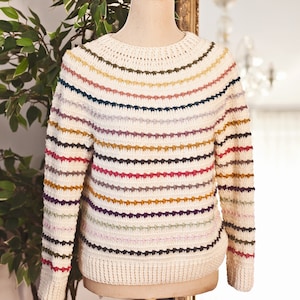 May include: Cream-colored sweater with horizontal stripes in various colors, including red, purple, and green. The sweater is displayed on a mannequin and has a round neckline and long sleeves. The knitwear is a cozy, handmade garment.