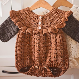 Könnte beinhalten: Ein handgefertigter, brauner Baby-Häkel-Strampler mit langen Ärmeln, einem Rüschenkragen und Knopfverschlüssen. Der Strampler wird auf einem Holzkleiderbügel präsentiert und zeigt das filigrane Lochmuster und die weiche Textur des Garns.