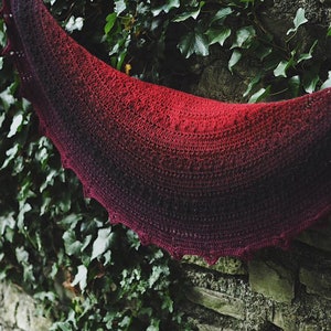 May include: A red and black ombre crochet shawl with a scalloped edge. The shawl is draped over a stone wall covered in green ivy.