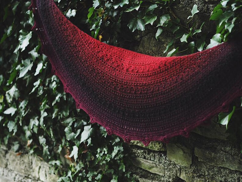 May include: A red and black ombre crochet shawl with a scalloped edge. The shawl is draped over a stone wall covered in green ivy.