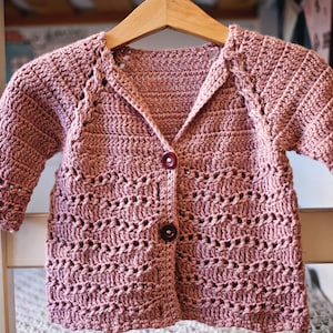 May include: A pink crocheted cardigan with a button closure. The cardigan has a simple, open stitch pattern and a slightly oversized fit.