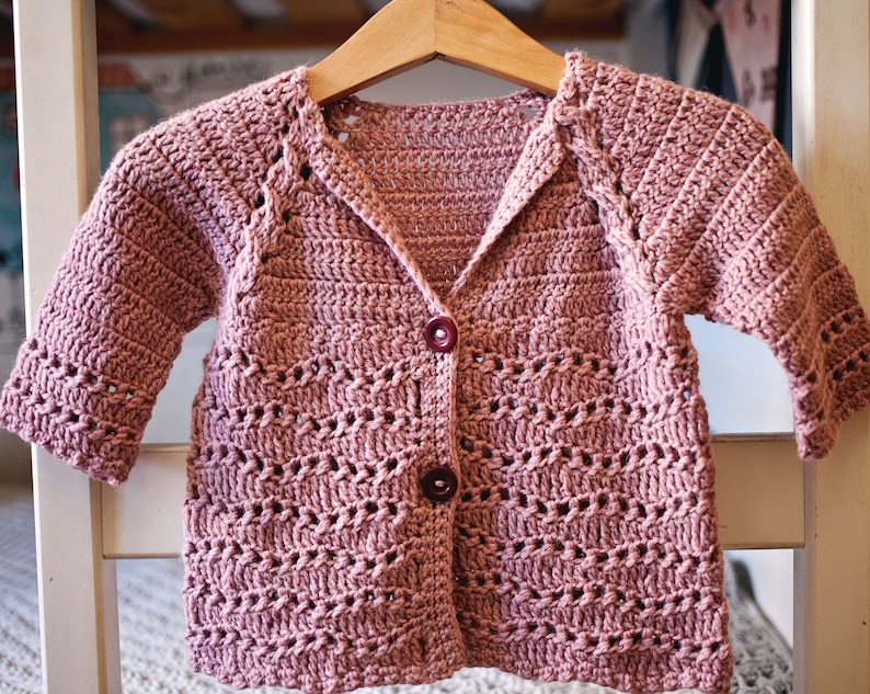 May include: A pink crocheted cardigan with a button closure. The cardigan has a simple, open stitch pattern and a slightly oversized fit.