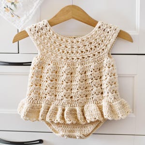 May include: A cream-coloured, handmade crochet romper with a ruffled hem, displayed on a wooden hanger. The romper features a detailed openwork design and is likely for a baby or toddler. The garment is made of soft yarn.