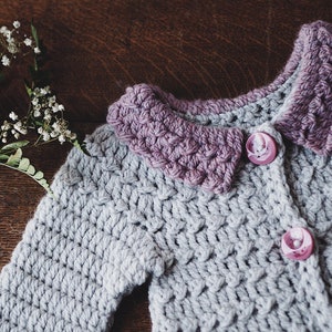 May include: A handmade crochet cardigan in light gray with a lavender collar and two pink buttons. The textured stitchwork and contrasting colors create a charming, cozy aesthetic. The cardigan is a soft, comfortable garment.
