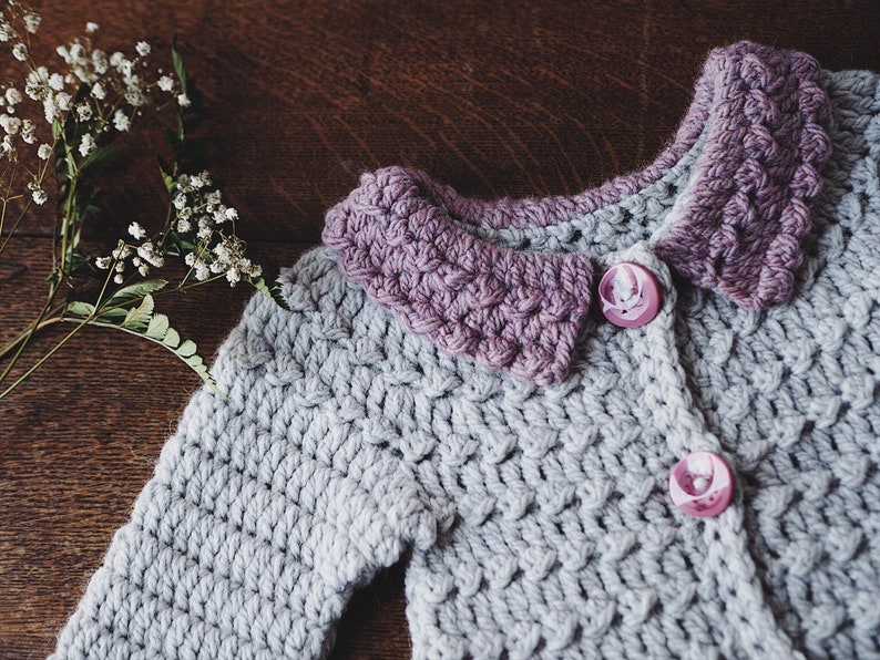 May include: A handmade crochet cardigan in light gray with a lavender collar and two pink buttons. The textured stitchwork and contrasting colors create a charming, cozy aesthetic. The cardigan is a soft, comfortable garment.