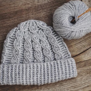 May include: A gray knitted beanie with a cable knit pattern and a folded brim. A ball of gray yarn and a knitting needle are also in the image.