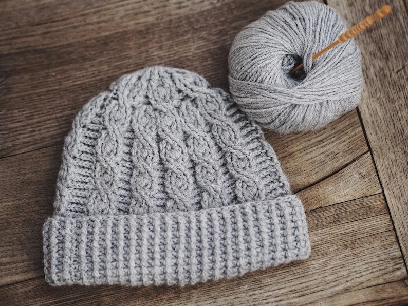 May include: A gray knitted beanie with a cable knit pattern and a folded brim. A ball of gray yarn and a knitting needle are also in the image.