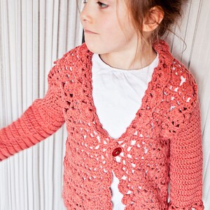 May include: A coral-colored, open-front crochet cardigan with a scalloped collar and sleeves. The handmade sweater features a single red button closure. The cardigan is worn over a white shirt.