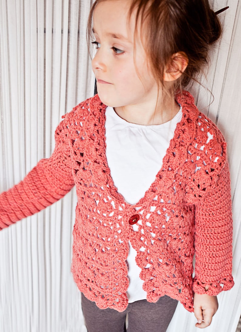 May include: A coral-colored, open-front crochet cardigan with a scalloped collar and sleeves. The handmade sweater features a single red button closure. The cardigan is worn over a white shirt.