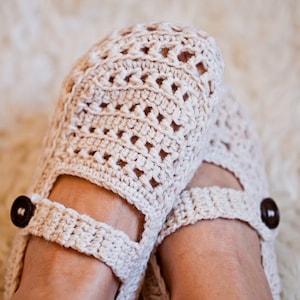 Puede incluir: Un par de zapatillas de crochet con un delicado diseño calado. Las zapatillas son de color crema y tienen un botón marrón a cada lado.