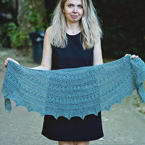 May include: A teal crochet shawl with a scalloped edge. The shawl is held by a person in front of a green background.