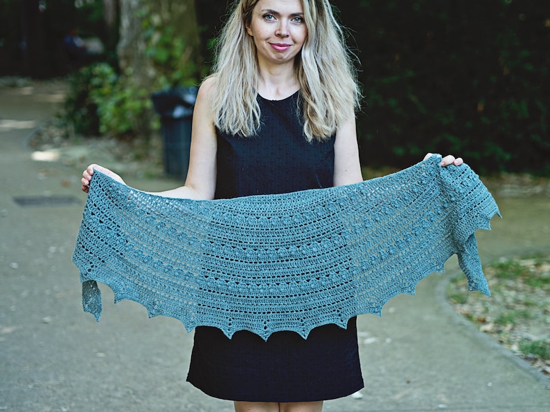 May include: A teal crochet shawl with a scalloped edge. The shawl is held by a person in front of a green background.