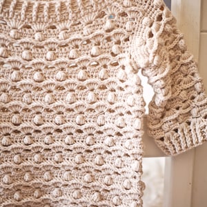 May include: A beige crocheted sweater with a textured, bobble stitch pattern. The sweater has a wide neckline and three-quarter sleeves.