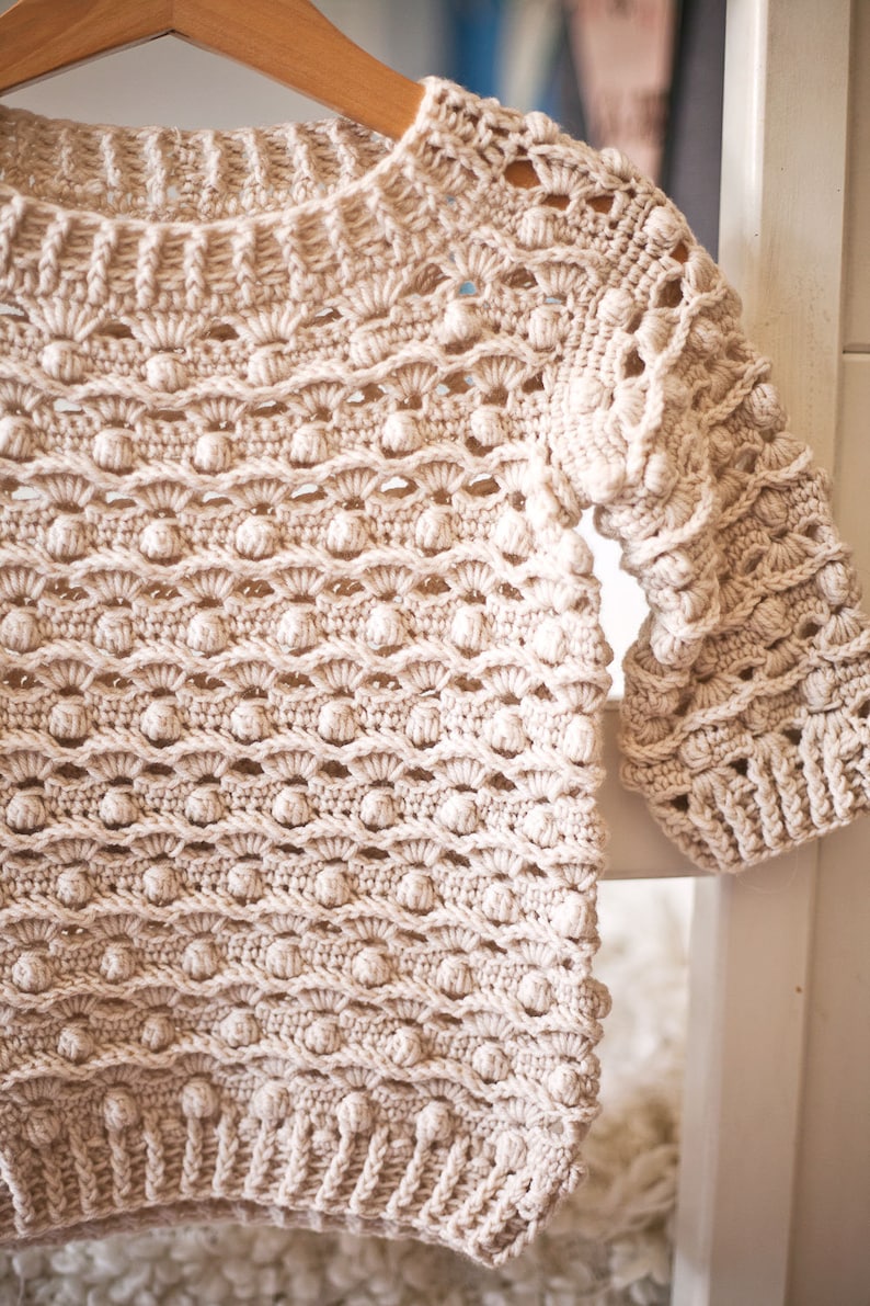 May include: A beige crocheted sweater with a textured, bobble stitch pattern. The sweater has a wide neckline and three-quarter sleeves.