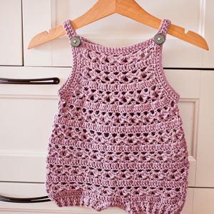 May include: A handmade, light purple crochet romper with a delicate openwork design. The romper features button closures on the straps and a scalloped edge. It is displayed on a wooden hanger.